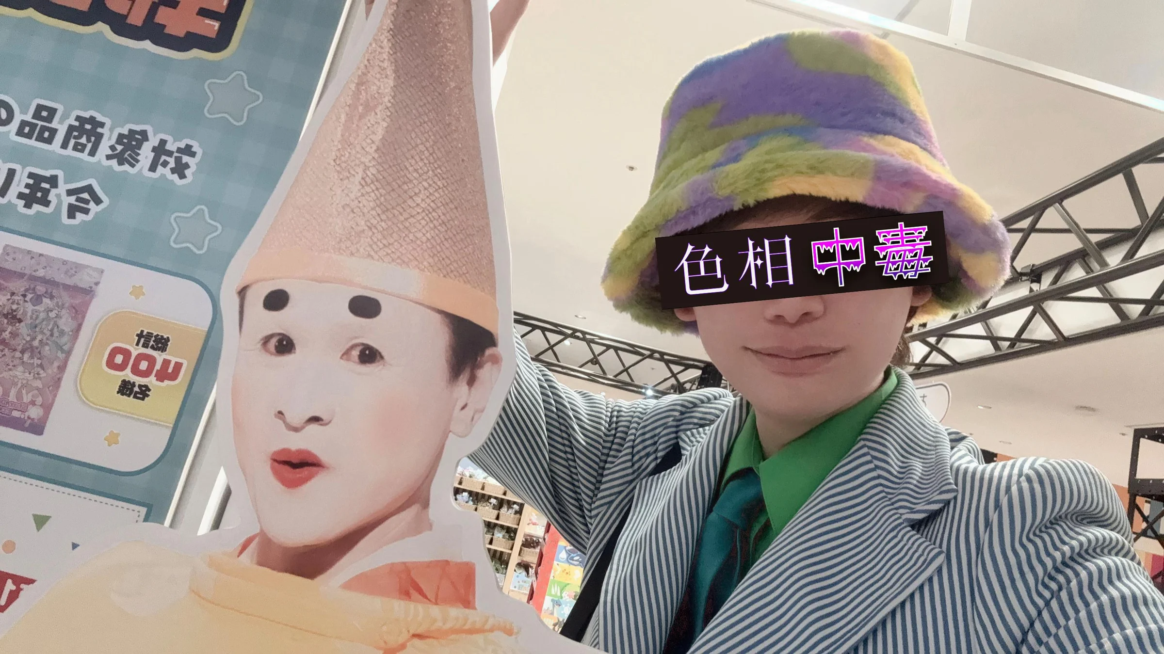 多色のふわふわ帽子と緑のシャツを着た色相中毒者が、パジャ麻呂の展示と並んだセルフィー